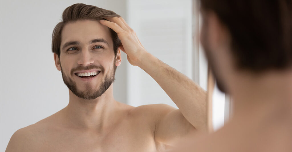 Shirtless man smiles in mirror as he runs hand through his hair.
